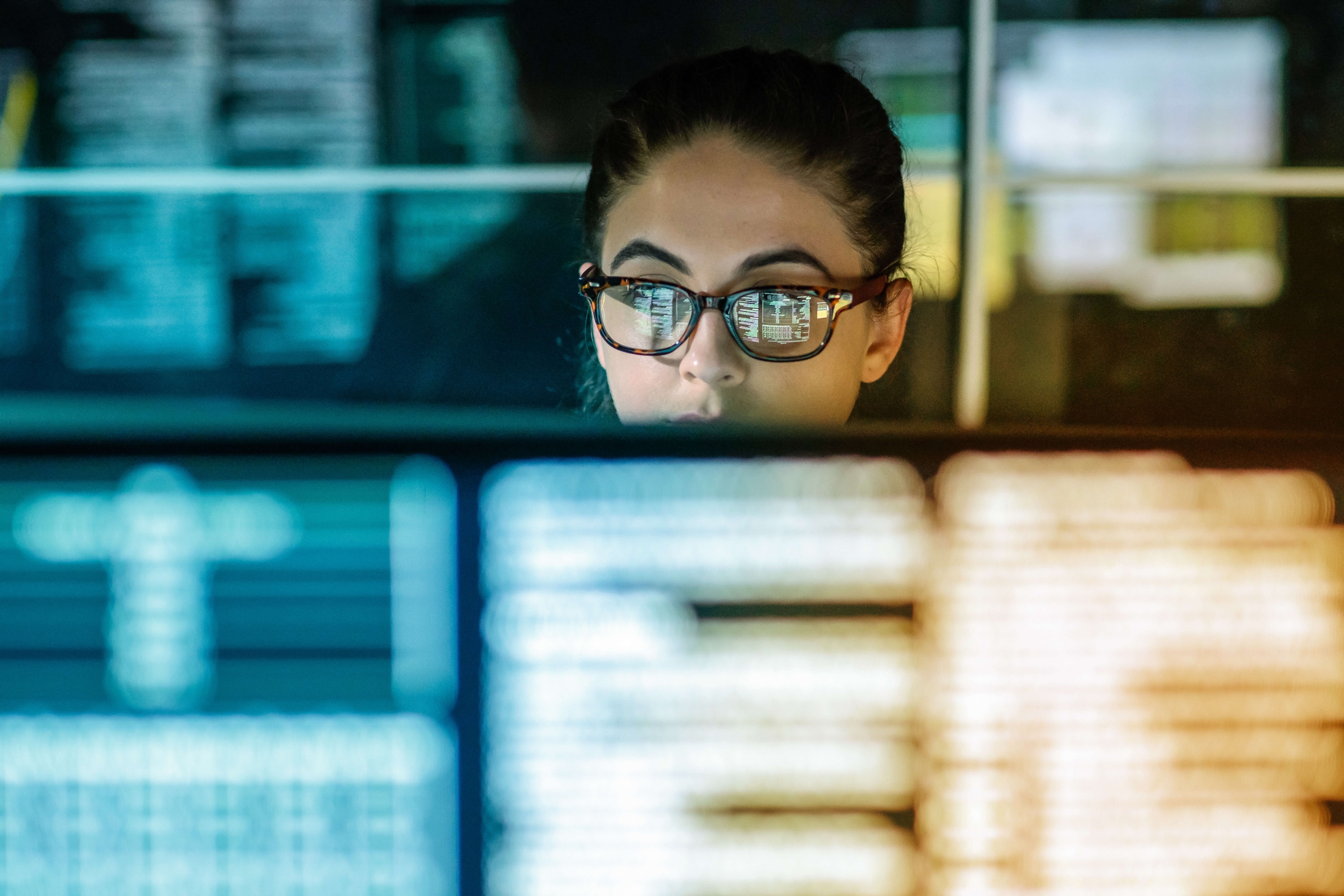 Frau mit Brille schaut in einem dunklen Raum in einen Bildschirm, vor und hinter ihr sieht man verschwommene Bildschirme.