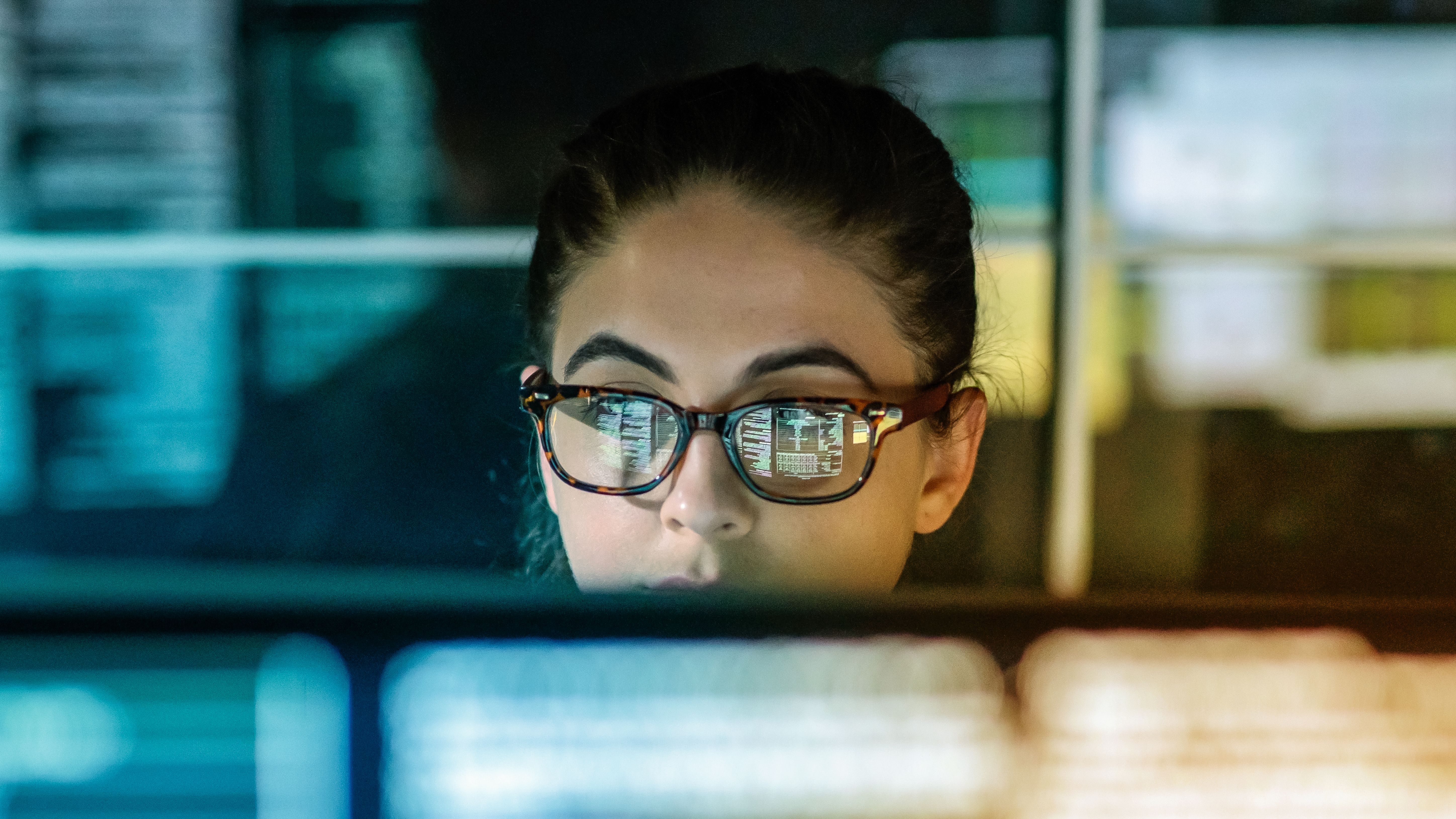 Frau mit Brille schaut in einem dunklen Raum in einen Bildschirm, vor und hinter ihr sieht man verschwommene Bildschirme.
