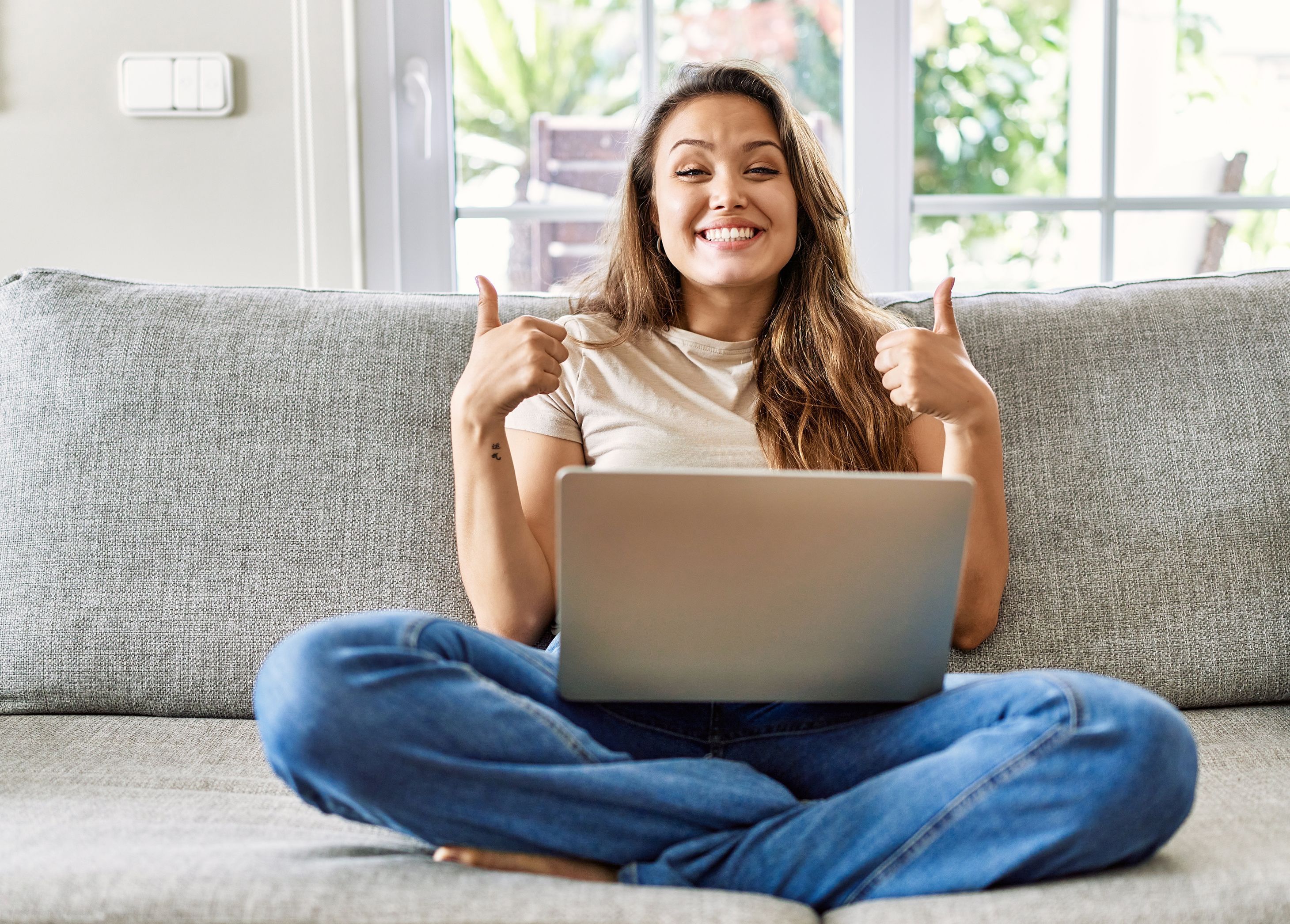 Junge Frau sitzt im Schneidersitz auf dem Sofa mit dem Laptop auf den Beinen und lächelt in die Kamera mit beiden Daumen nach oben