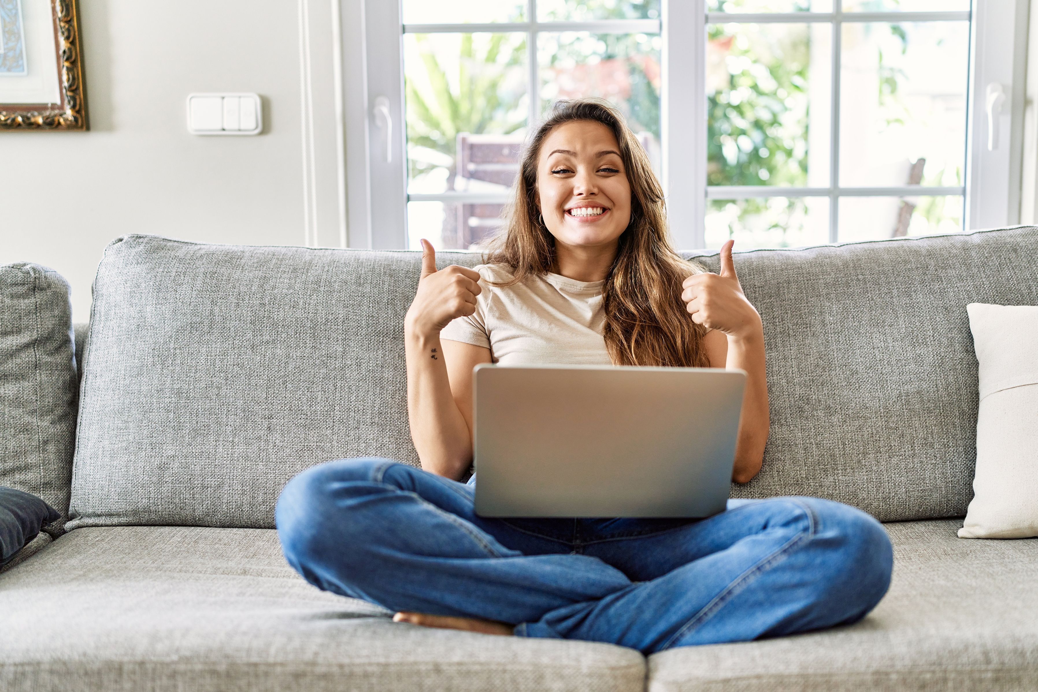 A young woman sits cross-legged on the sofa with a laptop on her lap, smiling at the camera with both thumbs up.