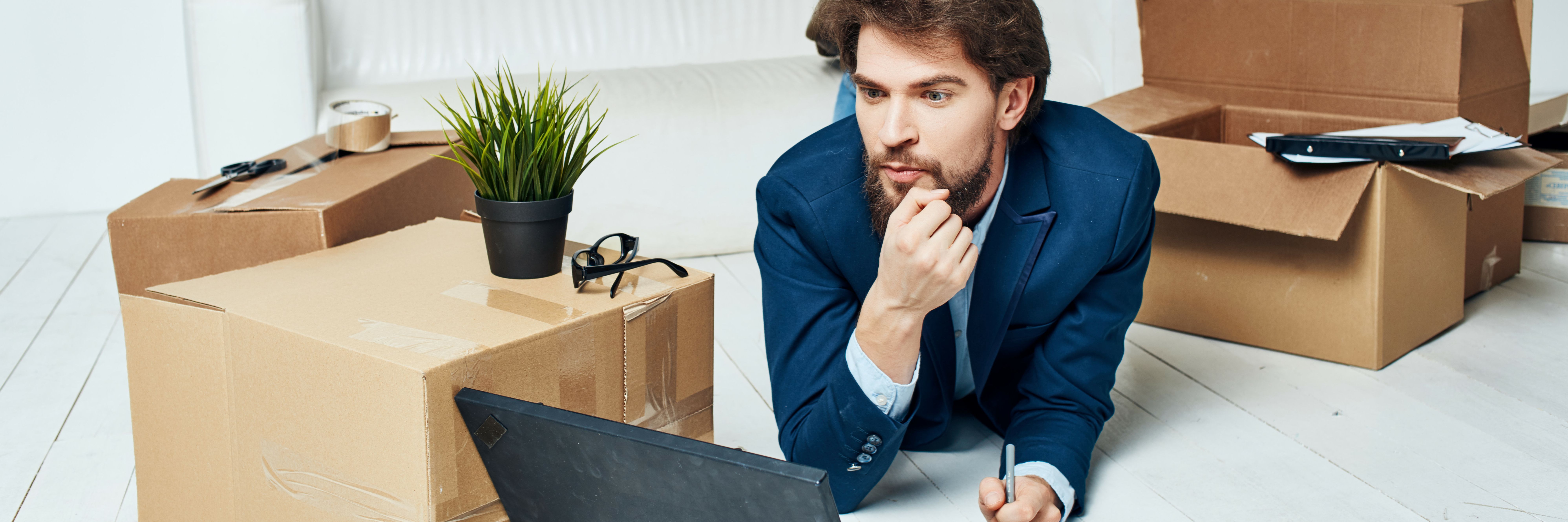 Mann liegt inmittem von Umzugskartons am Boden vor dem Laptop und überlegt sich, wie er seine Domain schnell und sicher zu Green transferiert.