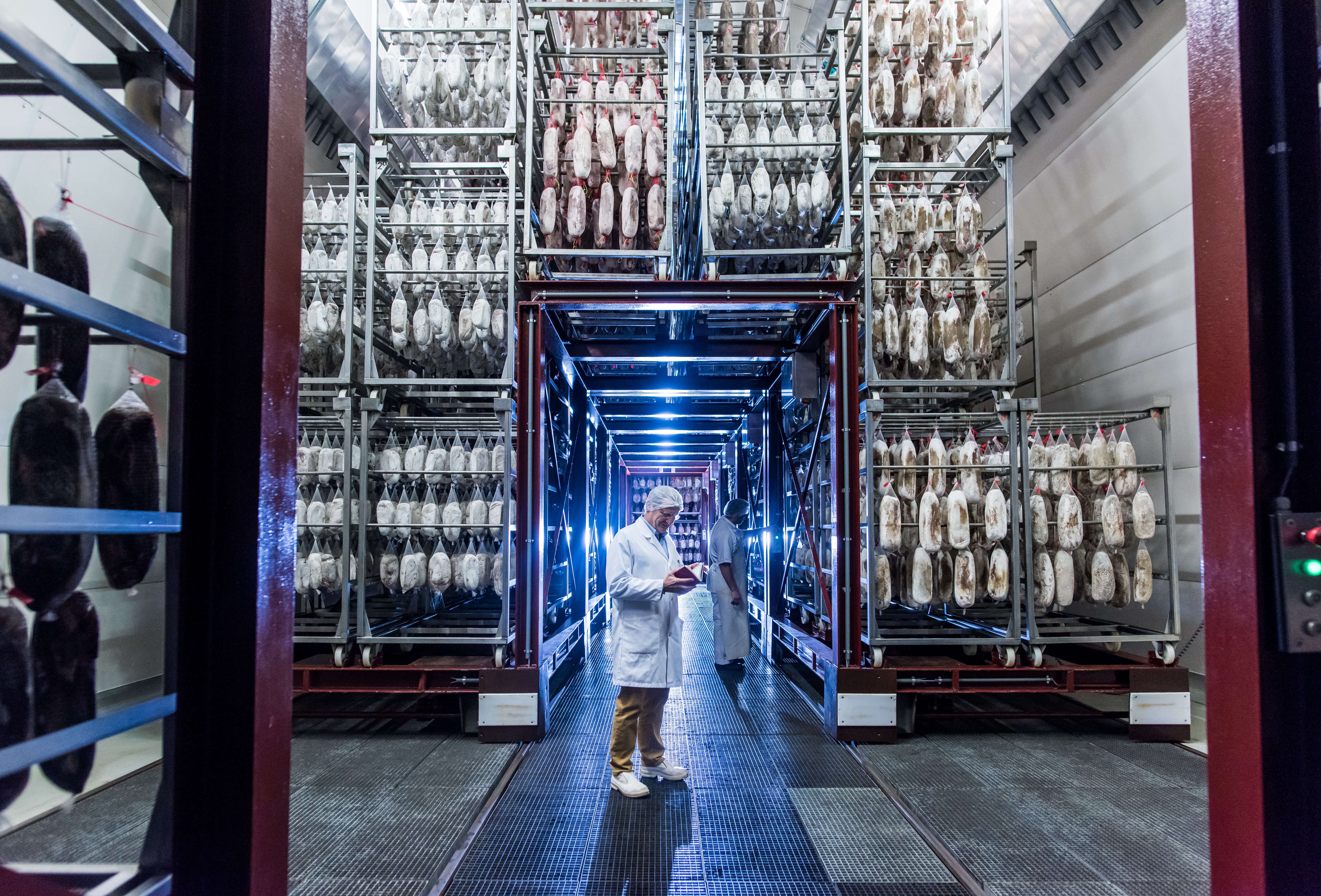 Zwei Männer mit weissen Kitteln, Schürzen und Hauben im Gang eines Kühlhaus mit vielen Regalen und hängenden Salamis. Einer schaut etwas in einem Notizbuch nach.